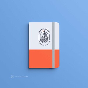 May include: A white and orange notebook with a black and white logo that says "Ready About Canvas Works" with a sailboat and a bird in the logo.