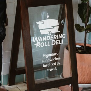 May include: A wooden A-frame sign with a glass front advertising "Wandering Roll Deli". The sign features a white silhouette of a food truck with a sandwich on top. The text "Signature sandwiches inspired by travel." is also on the sign.