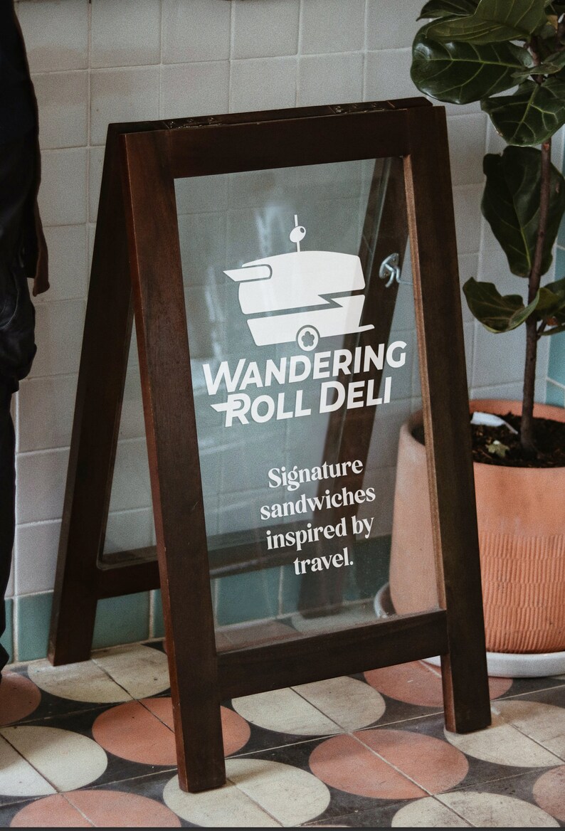 May include: A wooden A-frame sign with a glass front advertising "Wandering Roll Deli". The sign features a white silhouette of a food truck with a sandwich on top. The text "Signature sandwiches inspired by travel." is also on the sign.