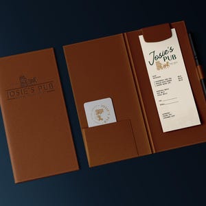 May include: A brown leather folder with a menu inside. The menu is for Josie's Pub and has a logo of a beer mug and a horse-drawn carriage. The folder has a pocket on the front for a business card.