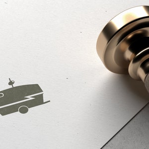 May include: A gold-toned metal stamp with a wooden handle. The stamp has a design of a vintage camper with a lightning bolt on the side. The stamp is on a white surface.