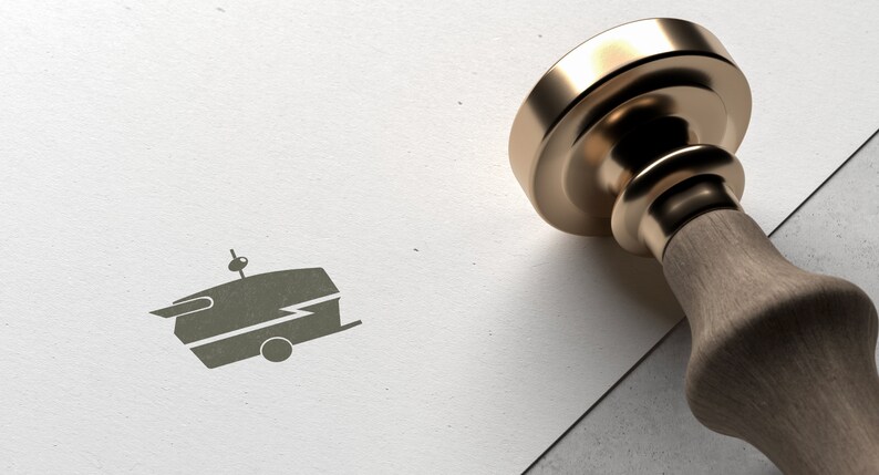 May include: A gold-toned metal stamp with a wooden handle. The stamp has a design of a vintage camper with a lightning bolt on the side. The stamp is on a white surface.