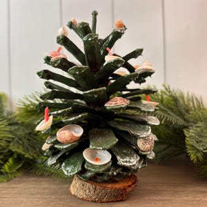 May include: A handcrafted Christmas tree made from a pine cone, painted dark green. The tree is decorated with small seashells and red accents. It is mounted on a small wooden base and set against a backdrop of greenery.