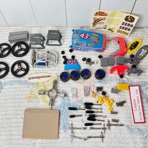 May include: A collection of model car parts, including wheels, chassis, and tools. The parts are in various colors, including blue, red, and black. Some parts have the number 43 on them. There are also stickers and a warning label.