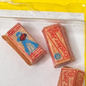 May include: Close-up of three miniature Cracker Jack boxes. The boxes are rectangular with red and tan packaging. One box features a blue cartoon figure. The words "Cracker Jack" are printed on the side of two boxes.