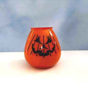 May include: Orange glass Halloween decoration shaped like a pumpkin with a black jack-o'-lantern face. The pumpkin has vertical black lines and a rounded shape. The background is a light blue.