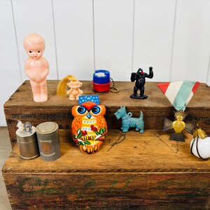 May include: A collection of vintage figurines and trinkets on a wooden box. Items include a pink doll, a blue dog, a gorilla, an owl, a medal, a perfume bottle, and two small metal containers. The items are arranged on a rustic wooden surface.