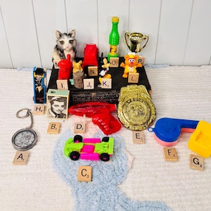 May include: A collection of vintage toys and collectibles, including a toy car, a red water gun, a gold trophy, a police badge, a cat figurine, and a keychain. The items are arranged on a white surface with scrabble tiles.