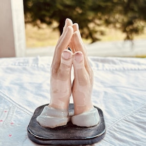 May include: A ceramic figurine depicting two hands pressed together in a prayer position. The hands are finished in a glossy, light peach-toned glaze, with pale blue cuffs at the wrists, all mounted on a smooth, dark, rectangular base.