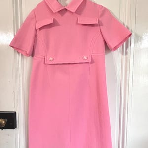 May include: A bright pink dress with short sleeves and a collared neckline. The dress features two small pockets on the chest and decorative buttons. The dress is hanging on a hanger against a white door.