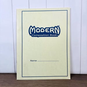 May include: A cream-colored composition book with a blue border. The cover features the word "MODERN" in a blue, stylized font above "Composition Book." Below is a line for the user to write their name.