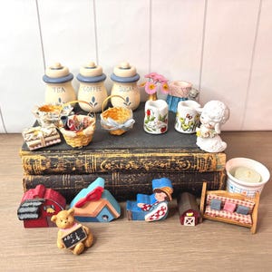 May include: A collection of miniature decorative items, including tea, coffee, and sugar canisters, baskets, floral vases, and figurines. The items are arranged on top of antique books and a wooden surface. A teddy bear holds a chalkboard with "ABC" written on it.