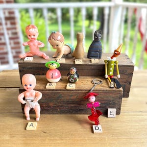 May include: A collection of vintage toys including two plastic baby dolls, a wooden bowling pin, a carved black cat, a wooden Pinocchio figure, a round pink toy, a small matryoshka doll, and a red beaded keychain, all labeled with letter tiles.