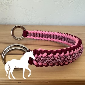 May include: A pink and burgundy braided horse collar with two silver metal rings. The collar is made of a tightly woven material and features a decorative pattern. A white silhouette of a horse is in the lower left corner.
