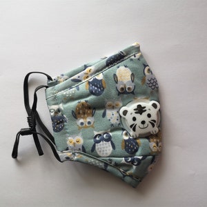 Pode incluir: Uma máscara facial azul clara com um padrão de corujas e um clipe branco em forma de tigre. A máscara tem alças elásticas pretas ajustáveis. O padrão de corujas inclui várias corujas.