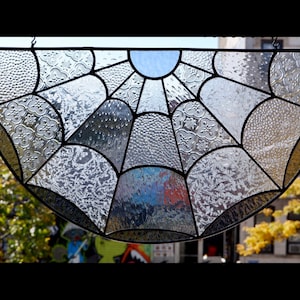 May include: A stained glass window in the shape of a half circle with a spiderweb design. The glass is clear with a variety of textures and patterns. The window is framed in black metal.