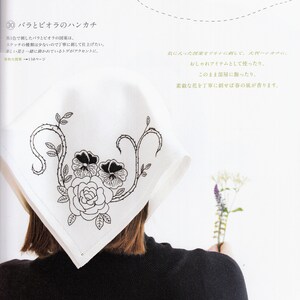 May include: A white handkerchief with black embroidery of a rose and two pansies. The handkerchief is folded and draped over the shoulder of a person wearing a black turtleneck sweater.