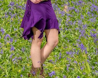 May include: A person wearing a purple wrap skirt with a ruffled hem, paired with brown boots. The skirt is tied at the waist with a drawstring. The background is a field of bluebell flowers.