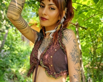 Peut inclure: Une femme portant un haut court en dentelle et en mesh marron avec une bordure verte. Le haut a un nœud à l'avant et est porté sur une jupe noire. La femme a des tatouages sur les bras et se tient devant un fond vert feuillu.