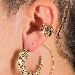May include: Gold ear cuff with an Om symbol design and a gold hoop earring with a feather design.