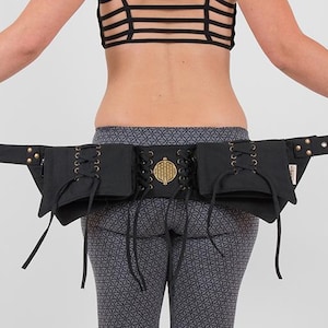 May include: Black belt bag with multiple pockets and a gold-colored buckle. The belt features a decorative gold emblem and is held open, showcasing its design. The bag is made of a dark material and has a long strap with multiple holes.