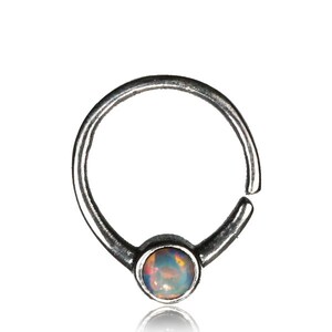 May include: Silver hoop nose ring with a small, round opal gemstone in the center. The opal is iridescent with shades of blue, green, and pink.