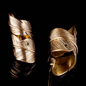 Op de afbeelding: Twee gouden ringen in de vorm van een veer. De ringen zijn gemaakt van metaal en hebben een getextureerd oppervlak. De ringen bevinden zich op een zwarte achtergrond.
