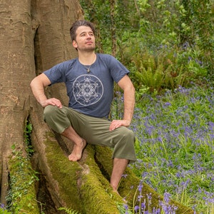 K&ouml;nnte beinhalten: Ein Mann in einem dunkelblauen T-Shirt mit einem wei&szlig;en geometrischen Muster sitzt auf einer Baumwurzel in einem Wald. Der Waldboden ist mit Blausternen bedeckt.