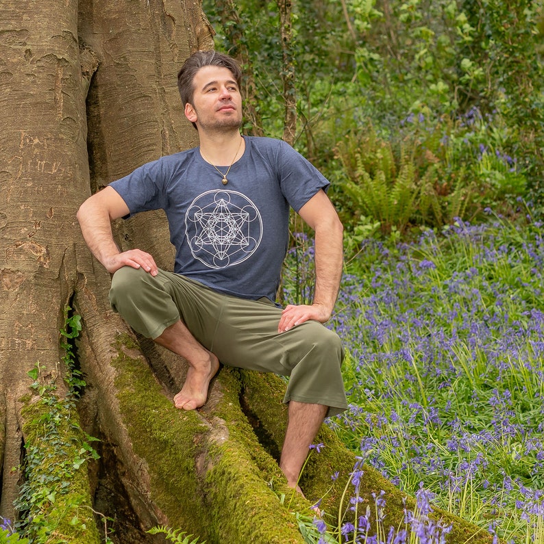 K&ouml;nnte beinhalten: Ein Mann in einem dunkelblauen T-Shirt mit einem wei&szlig;en geometrischen Muster sitzt auf einer Baumwurzel in einem Wald. Der Waldboden ist mit Blausternen bedeckt.