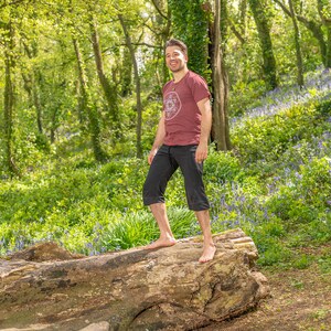 K&ouml;nnte beinhalten: Ein Mann in einem maroonfarbenen T-Shirt mit einem wei&szlig;en Grafikdesign steht auf einem gro&szlig;en Baumstamm in einem Wald. Der Waldboden ist mit Blausternen bedeckt.