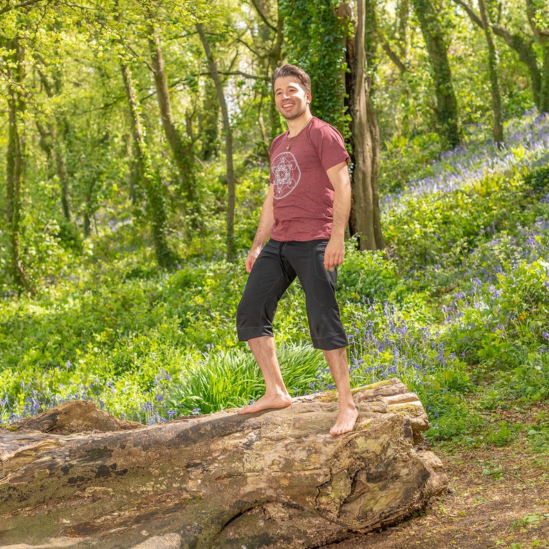 K&ouml;nnte beinhalten: Ein Mann in einem maroonfarbenen T-Shirt mit einem wei&szlig;en Grafikdesign steht auf einem gro&szlig;en Baumstamm in einem Wald. Der Waldboden ist mit Blausternen bedeckt.