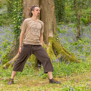 K&ouml;nnte beinhalten: Eine Person tr&auml;gt ein beiges T-Shirt mit einem geometrischen Muster und eine braune, kurze Hose und steht in einer Waldlandschaft. Die Person steht in der N&auml;he eines Baumes mit Moos, das an den Wurzeln w&auml;chst. Der Waldboden ist mit blauen Wildblumen bedeckt.