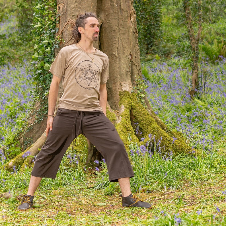 K&ouml;nnte beinhalten: Eine Person tr&auml;gt ein beiges T-Shirt mit einem geometrischen Muster und eine braune, kurze Hose und steht in einer Waldlandschaft. Die Person steht in der N&auml;he eines Baumes mit Moos, das an den Wurzeln w&auml;chst. Der Waldboden ist mit blauen Wildblumen bedeckt.