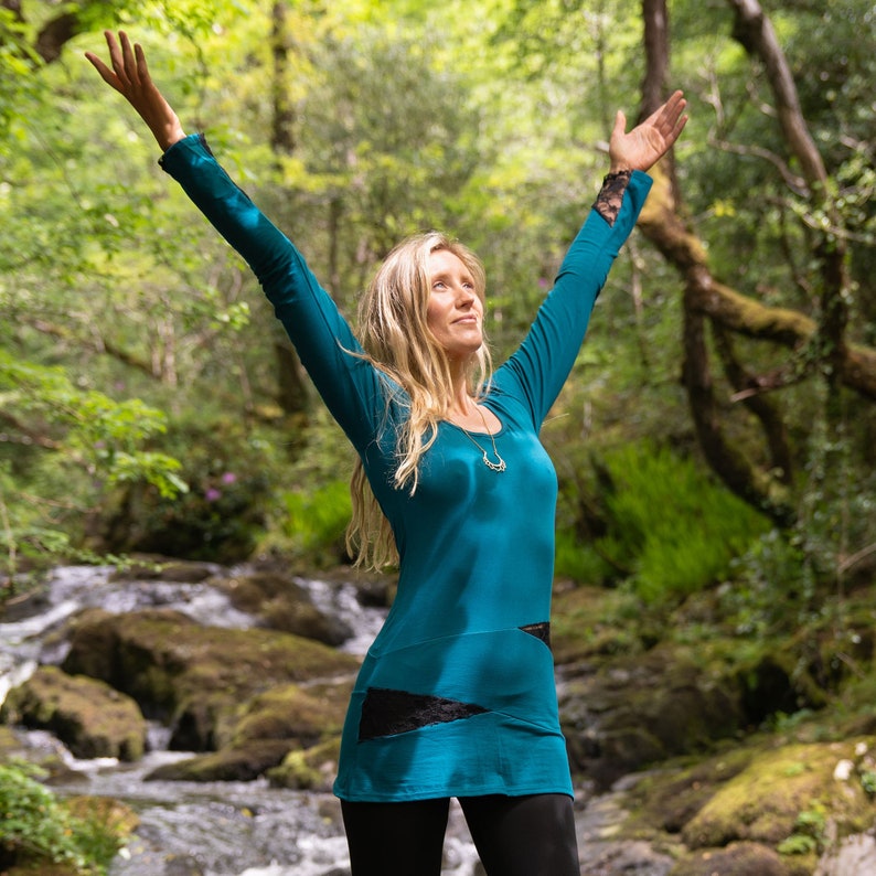 May include: A woman wearing a teal long-sleeved shirt with black accents and black leggings, standing with her arms raised in a forest setting.