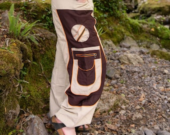 May include: Beige and brown pants with a unique design. The pants feature a large brown panel on the side with a circular accent and a pocket with an orange zipper. The pants are worn with black sandals.