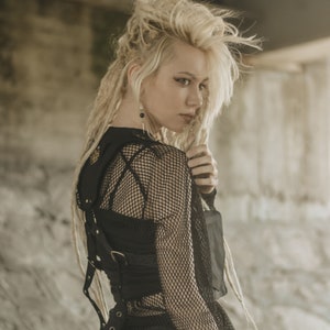 May include: A woman with blonde dreadlocks wears a black mesh top with a leather harness. The harness has straps and buckles. The woman is looking over her shoulder.