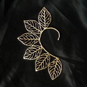 May include: Gold leaf ear cuff with a delicate, intricate design. The cuff is made of metal and has a curved shape that fits comfortably around the ear.