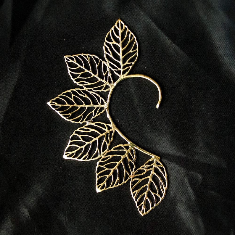 May include: Gold leaf ear cuff with a delicate, intricate design. The cuff is made of metal and has a curved shape that fits comfortably around the ear.