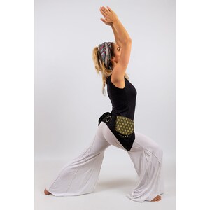 Op de afbeelding: Een vrouw die een witte broek en een zwart tanktop draagt met een zwarte en gouden gepatroneerde riemtas. Ze beoefent yoga met haar armen boven haar hoofd.