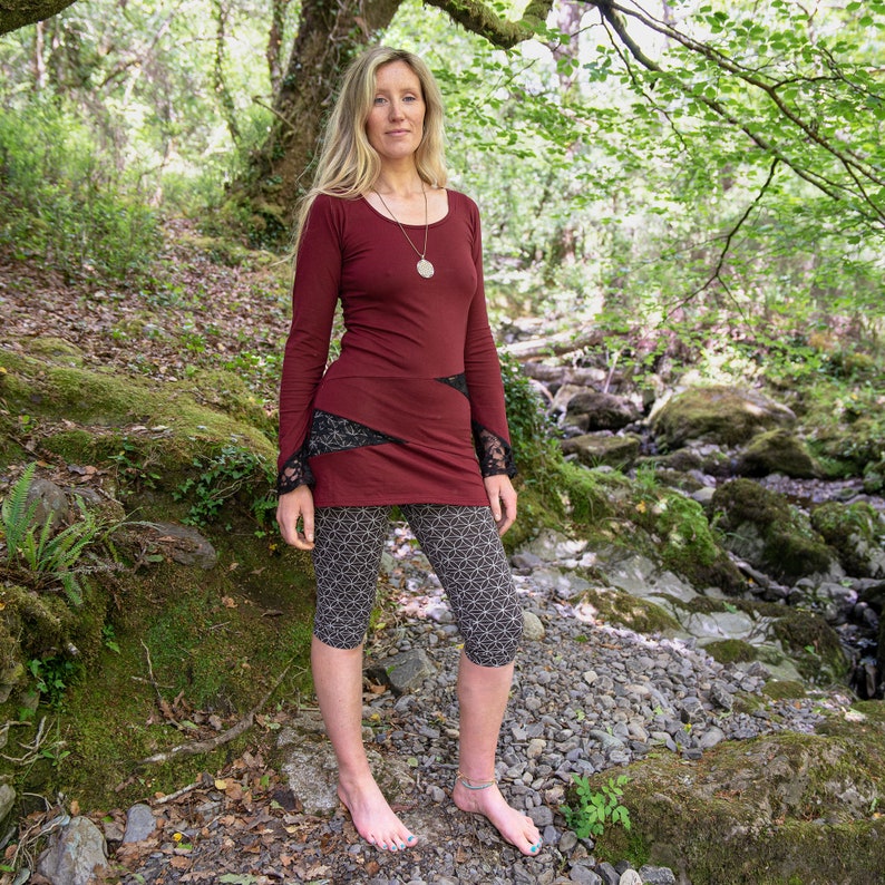 May include: A woman wearing a maroon long-sleeved top with black lace detailing on the sleeves and a geometric pattern on the leggings. She is standing in a forest setting.