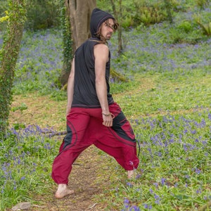 Puede incluir: Una persona con una camisa negra con capucha y pantalones rojos con dise&ntilde;os negros se encuentra en un entorno boscoso. La persona est&aacute; de espaldas a la c&aacute;mara y mirando por encima del hombro. El suelo del bosque est&aacute; cubierto de flores azules.