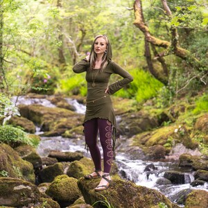 May include: A woman wearing a green hooded top with a long, flowing skirt and purple leggings with a floral design. She is standing on a rock by a stream in a forest.