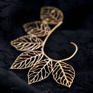 May include: Gold leaf ear cuff with a delicate, intricate design. The cuff is made of metal and features a series of leaves that are connected to a curved wire.
