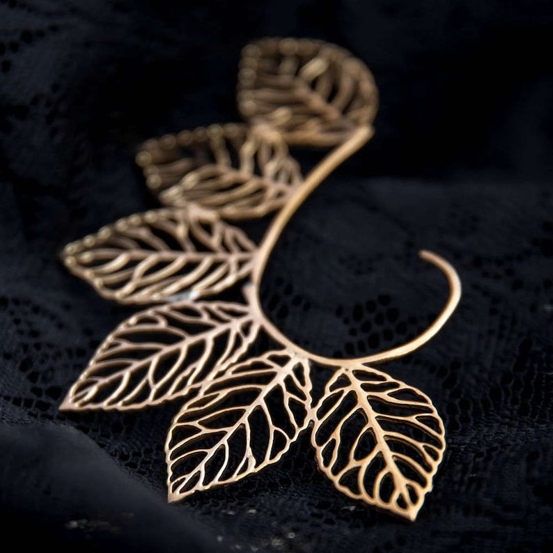 May include: Gold leaf ear cuff with a delicate, intricate design. The cuff is made of metal and features a series of leaves that are connected to a curved wire.