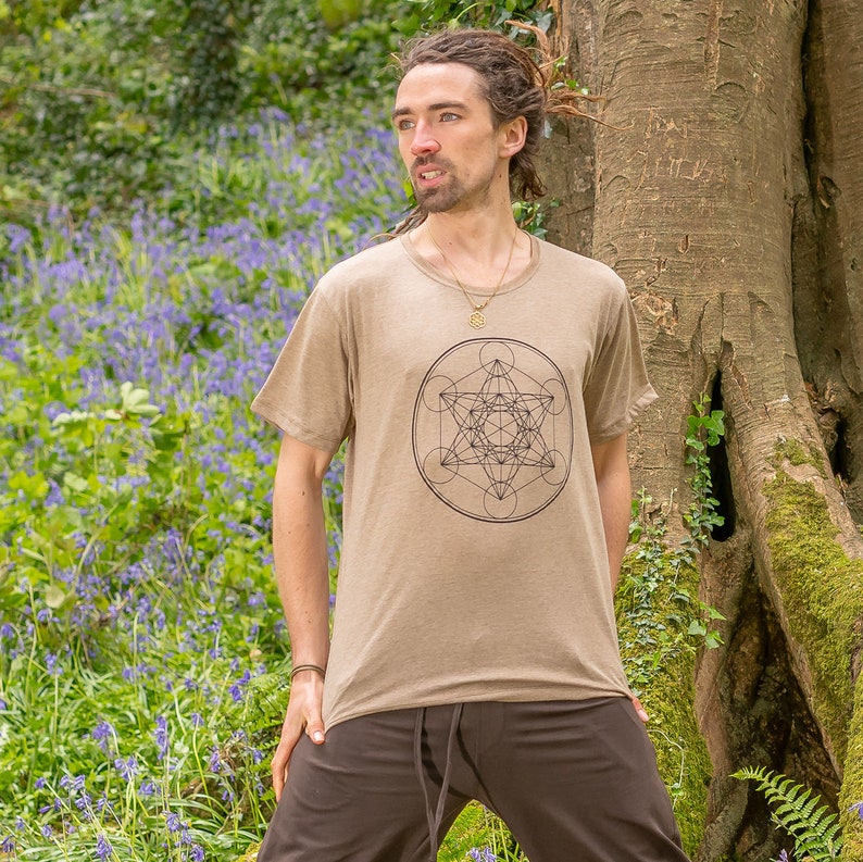 K&ouml;nnte beinhalten: Ein beiges T-Shirt mit einem schwarzen geometrischen Design auf der Vorderseite. Das Design ist ein Kreis mit einem Stern im Inneren und anderen geometrischen Formen darum herum.