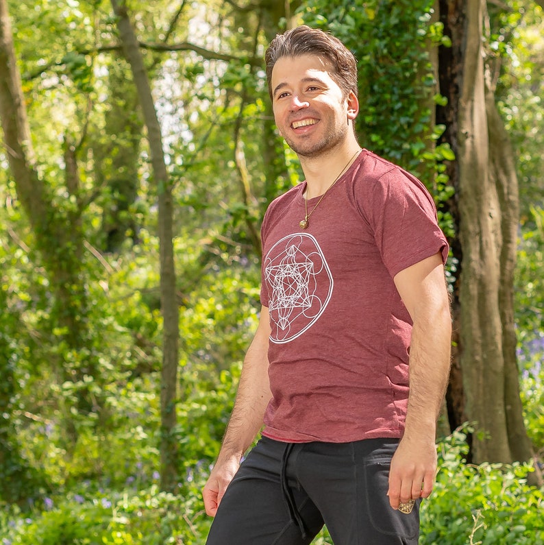 K&ouml;nnte beinhalten: Ein Mann tr&auml;gt ein weinrotes T-Shirt mit einem wei&szlig;en geometrischen Muster und eine schwarze Hose. Er steht in einem Wald mit gr&uuml;nen B&auml;umen und blauen Blumen.
