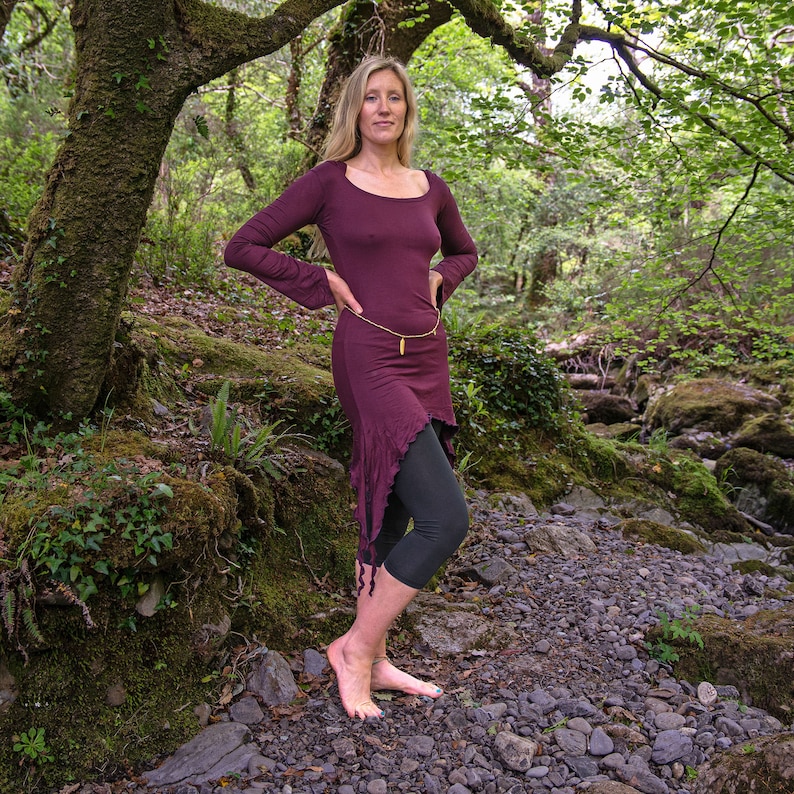 May include: A woman wearing a long, burgundy dress with a gold chain belt and black leggings. She is standing in a forest setting with green foliage and rocks.