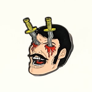 May include: A black and white enamel pin depicting a man's face with two yellow knives sticking through his eyes. Blood is dripping from the wounds. The man has a mustache and is screaming with his mouth open.