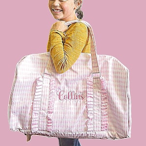 May include: A pink and white gingham travel bag with the name "Collins" embroidered in pink. The bag has pink ruffle accents and a matching strap. The bag is being carried by a person.