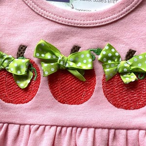 May include: Pink baby dress with three embroidered red apples. Each apple has a brown stem and a green polka dot bow. The dress has a ruffled hemline and a round neckline.